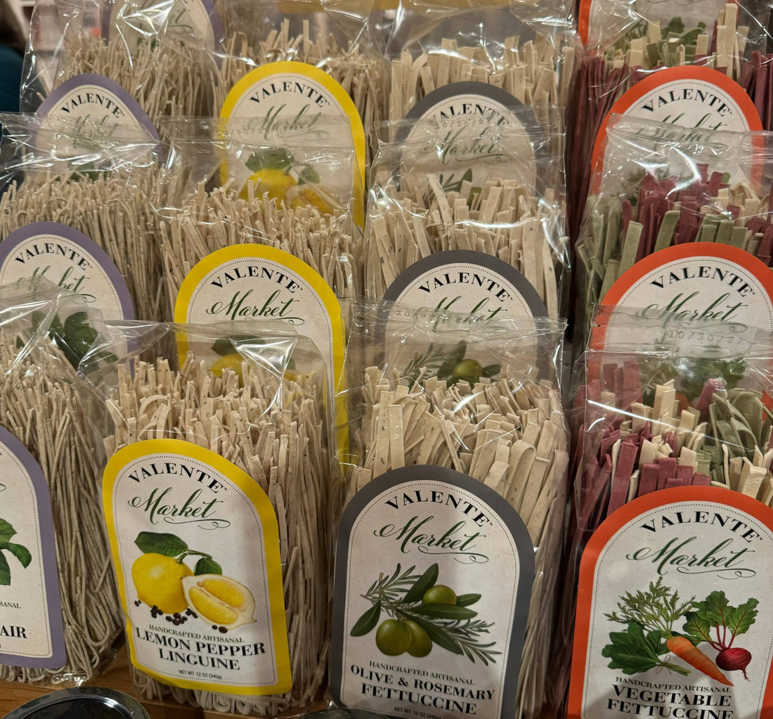 Assorted Valente Market artisanal pasta flavors in clear packaging on display, including lemon pepper linguine, olive and rosemary fettuccine, and vegetable fettuccine.