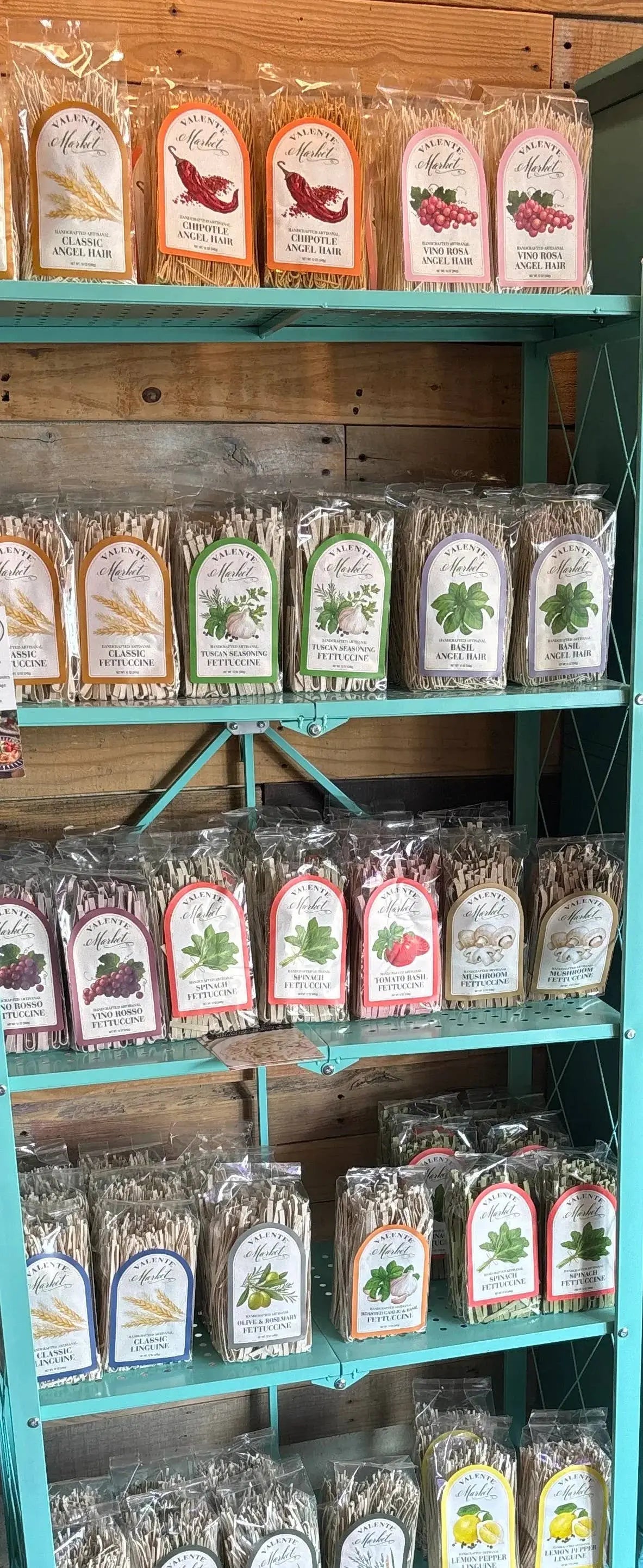 Assorted gourmet pasta varieties in clear packaging on teal metal shelves, rustic wood background.