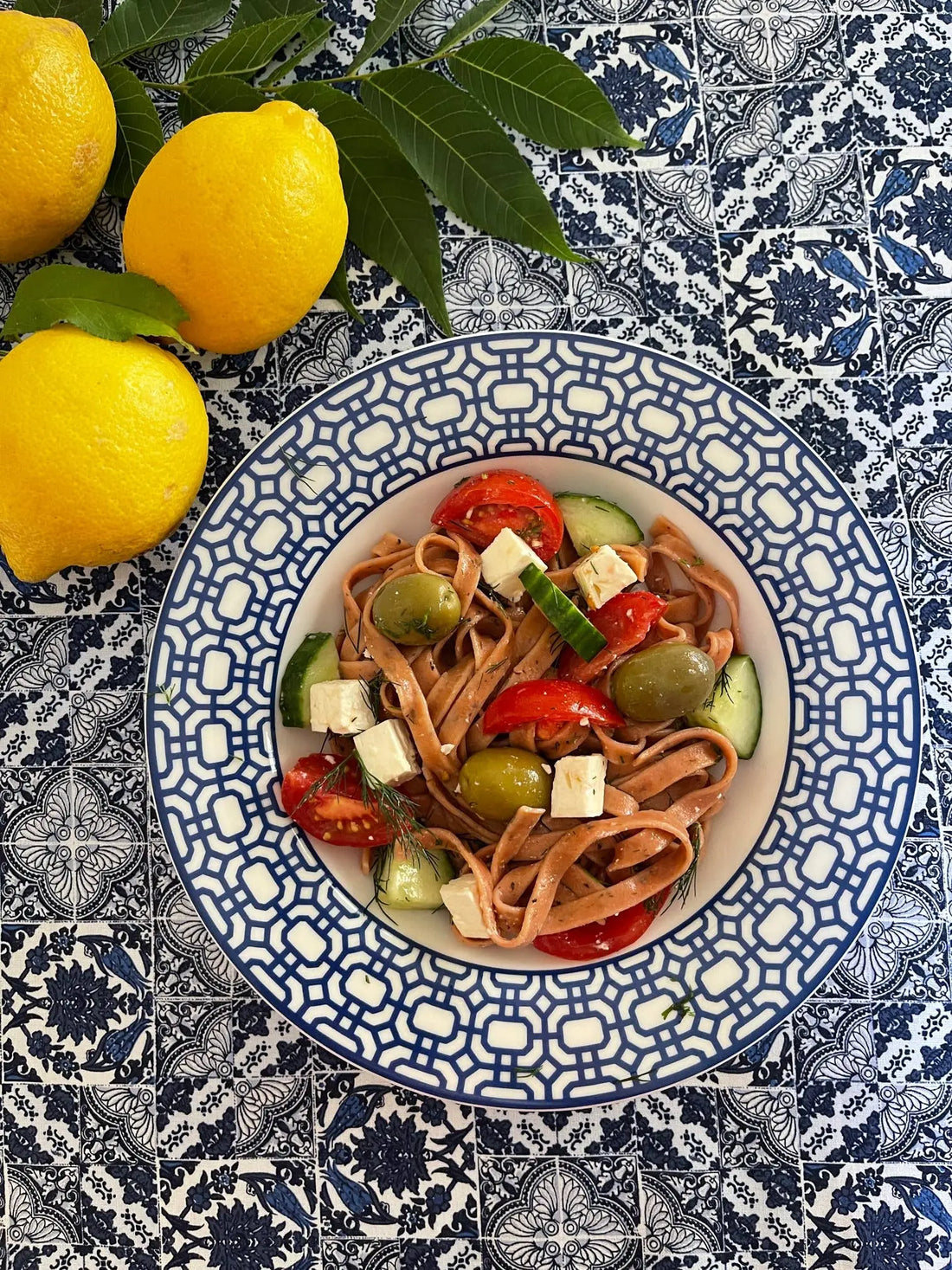 Mediterranean pasta salad with olives, feta, tomato, cucumber on patterned plate beside fresh lemons