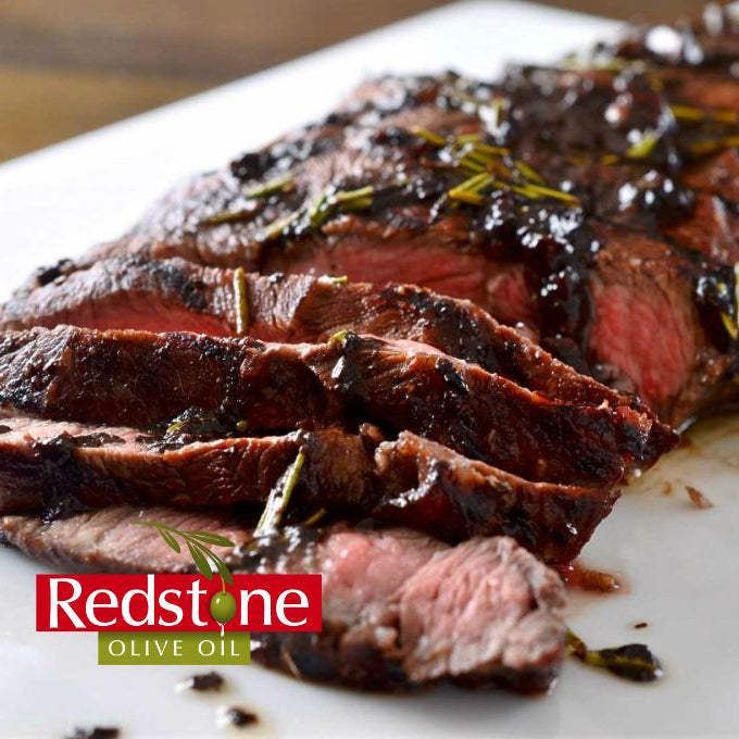 Sliced steak with rosemary on a white plate, featuring Redstone Olive Oil branding.