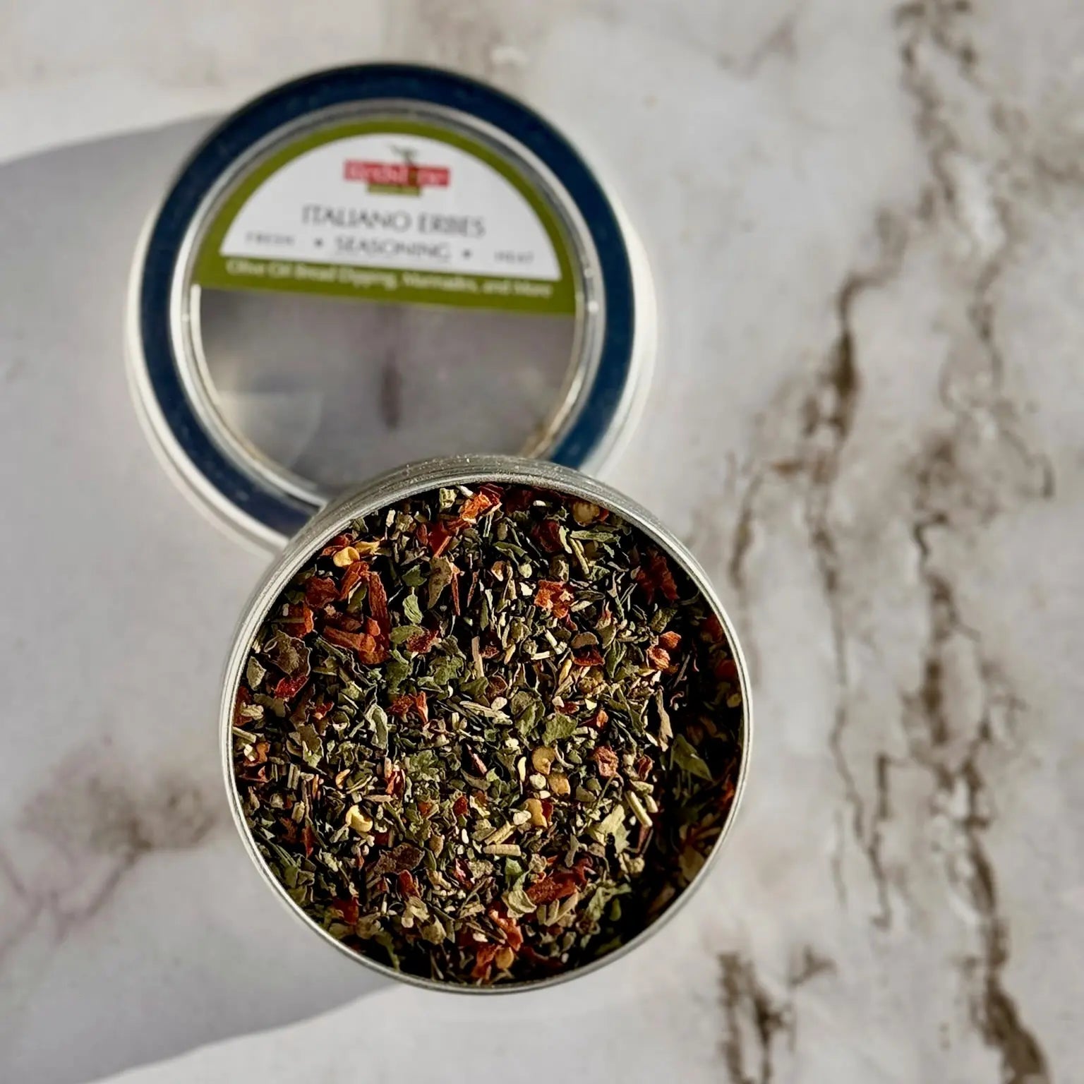 Open tin of Italiano herbs seasoning blend with colorful dried herbs on marble surface.