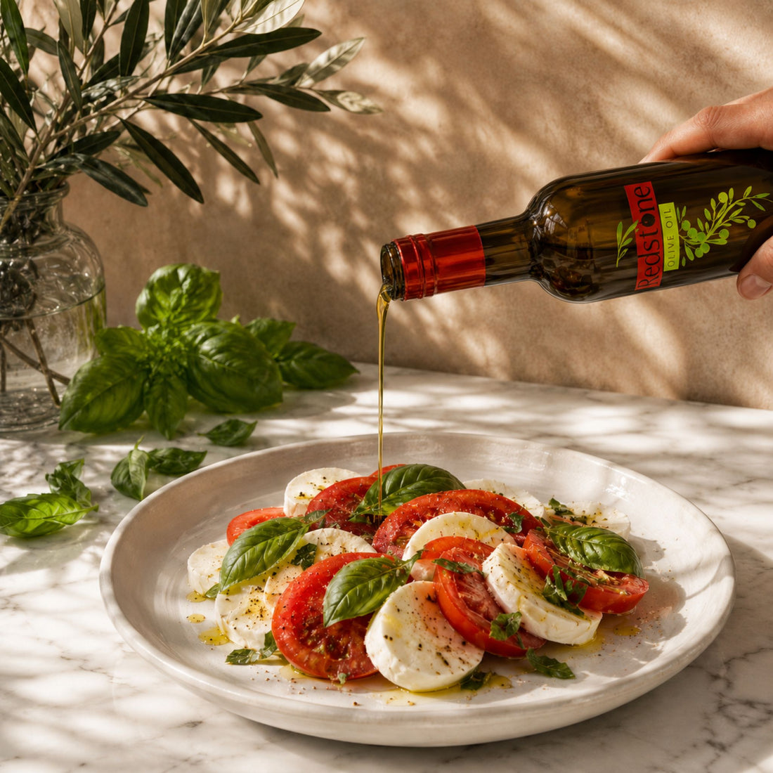 Hand pouring Redstone Olive Oil Basil Infused Olive Oil over a classic caprese salad of sliced heirloom tomatoes, fresh mozzarella, and whole basil leaves on a white ceramic plate, with fresh basil sprigs and an olive branch in a glass vase in the background on a marble surface