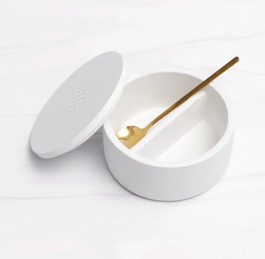 Ceramic salt cellar with white lid and gold spoon on a marble surface