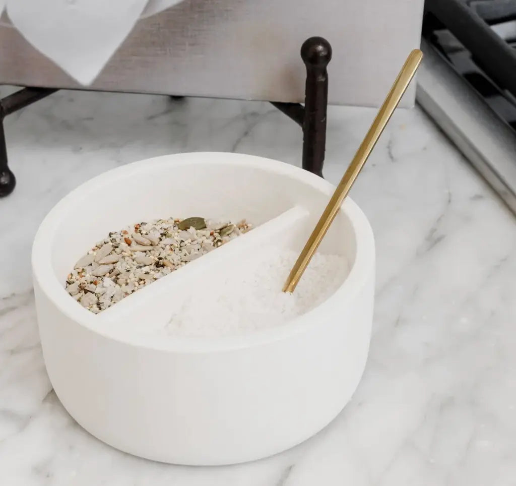 White salt cellar with gold spoon, holding salt and seasoning blend on marble kitchen counter