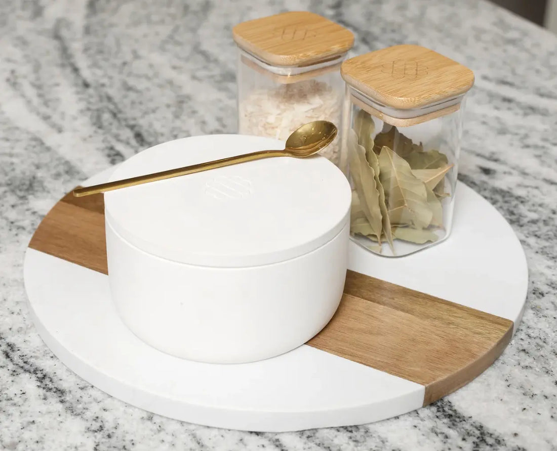 Ceramic salt cellar with gold spoon, spice jars, and bay leaves on a marble countertop