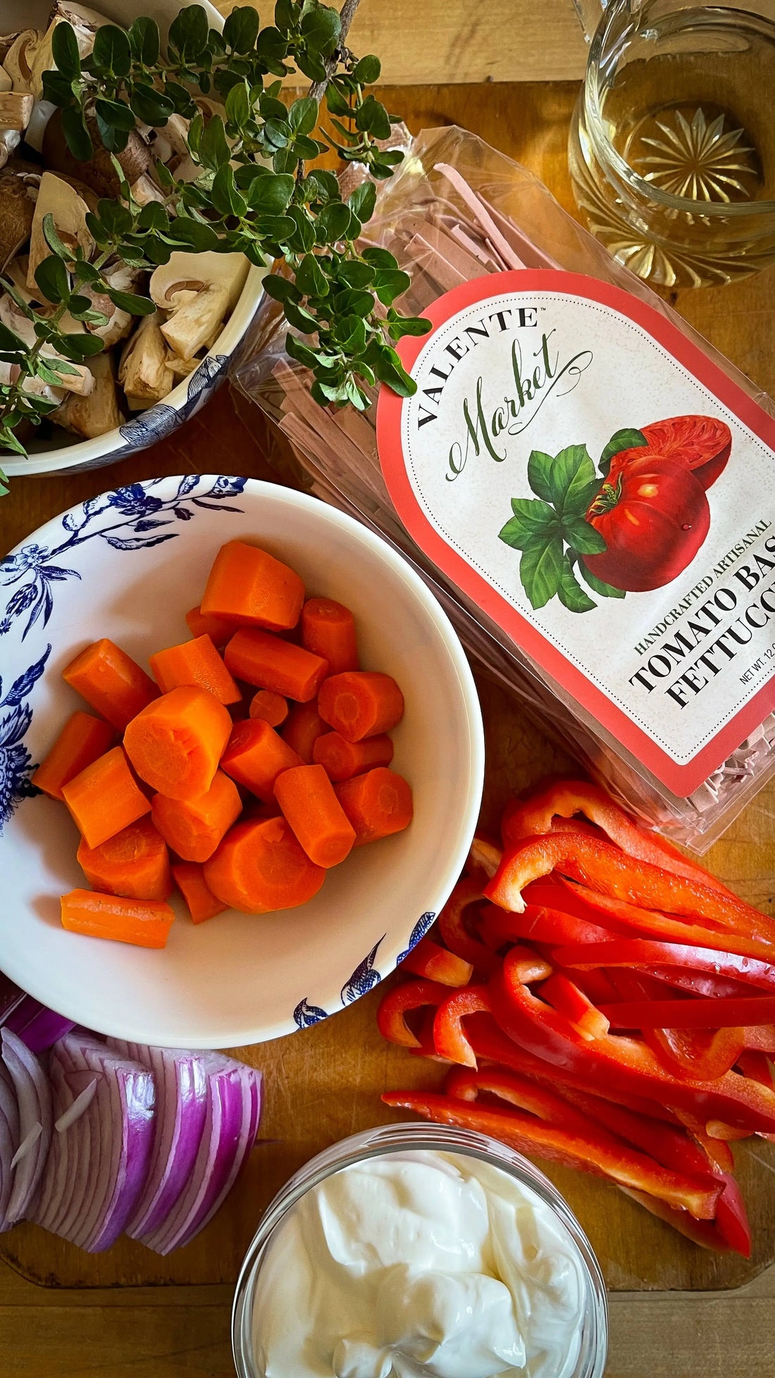 Tomato basil fettuccine, fresh herbs, carrots, red bell pepper, mushrooms, onion, sour cream, and white wine on a wooden surface.