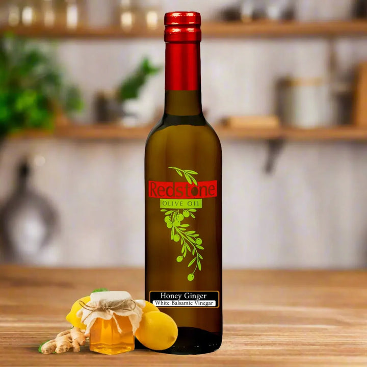 Bottle of Redstone Honey Ginger White Balsamic Vinegar with a honey jar, lemons, and ginger root on a kitchen counter