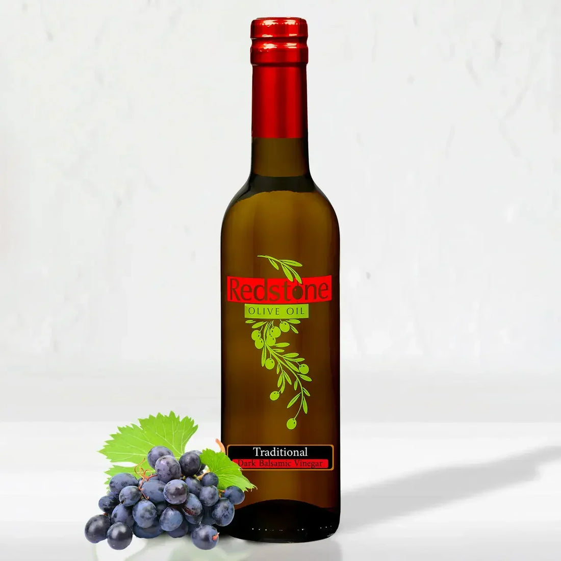 A tall glass bottle of Redstone Olive Oil brand Traditional Dark Balsamic Vinegar with a red foil cap capsule, displayed next to a cluster of dark purple grapes.
