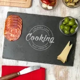 Slate cheese board with sliced salami, green olives, cheese wedges, and engraved cooking text